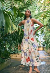 Abito lungo floreale con design a collo alto, caratterizzato da colori vivaci, strati sovrapposti e un tessuto leggero, ambientato su uno sfondo tropicale.