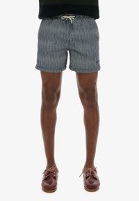 Bañador a rayas en azul marino con cintura elástica y cordón. Usado con zapatos de barco de cuero marrón. Los pantalones cortos cuentan con bolsillos laterales.