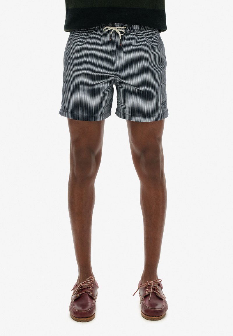 Bañador a rayas en azul marino con cintura elástica y cordón. Usado con zapatos de barco de cuero marrón. Los pantalones cortos cuentan con bolsillos laterales.