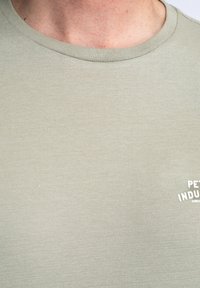 Camiseta de algodón verde claro con cuello redondo acanalado, que presenta un acabado texturizado y un pequeño logo blanco impreso en la parte inferior derecha.