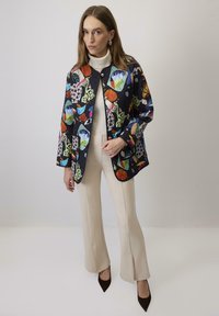 Giacca trapuntata nera con motivi geometrici e floreali colorati, dotata di bottoni a pressione, maniche lunghe e una vestibilità comoda. Abbinata a pantaloni beige a palazzo.
