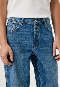 Blå denimjeans med høy midje, fem-lommers design og synlige sømmer. Stoffet har en litt falmet tekstur og finish.