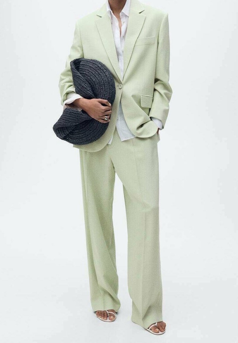 Personne portant un costume oversize vert clair avec une chemise blanche, tenant un grand objet tricoté gris foncé et portant des sandales blanches à bout ouvert.