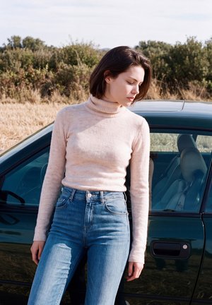 Une femme en pull clair et en jean se tient à côté d’une voiture sombre dans un champ sec bordé d’arbustes.