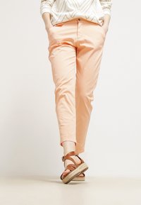Jasno brązowe bawełniane spodnie o slim fit, z kieszeniami bocznymi i podwiniętym dołem. Połączone z brązowymi sandałami na paski na neutralnej powierzchni.