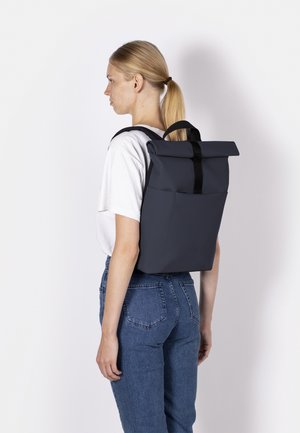 Woman with blonde ponytail wearing white t-shirt and blue jeans, carrying a large black roll-top backpack on her back, facing sideways.