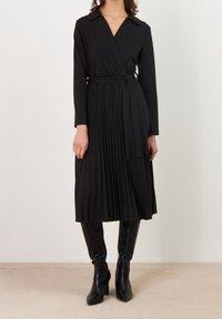 Černé plisované šaty s překládaným designem, dlouhými rukávy a límcem. Obsahují ladící pásek, nošené přes černé boty ke kolenům.