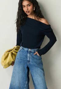 Navy off-shoulder långärmad topp i kombination med högmidjade ljusblå vida jeans. Modellen håller en gul samlad handväska.