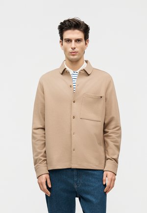 Νέος άνδρας με σκούρα μαλλιά, που φοράει μπεζ κουμπωτό σακάκι πάνω από λευκό και μπλε ριγέ πουκάμισο, μπλε τζιν, στέκεται μπροστά σε μονόχρωμο φόντο.