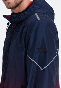 Marineblauwe waterdichte jas met een capuchon, rode accenten, reflecterende strips op de schouders en een logo patch op de mouw. Gladde textuur.