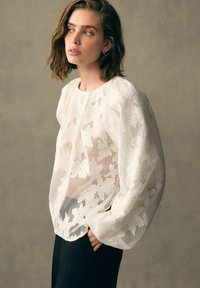 Blouse en dentelle blanche avec un motif floral, texture transparente et manches ballon. Associée à un pantalon noir, le design présente un col rond.