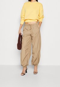 Sudadera amarilla recortada con el logo de Nike, combinada con pantalones sueltos beige con puños elásticos, y un bolso texturizado marrón. Sandalias de tacón negras.