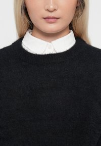 Schwarzer Strickpullover mit rundem Ausschnitt und strukturiertem Finish. Darunter trägt man ein weißes Hemd mit Kragen und Knopfdetails.