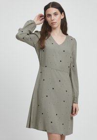 Khakifarbenes Kleid mit V-Ausschnitt und langen Puffärmeln, versehen mit schwarzen Punkten. Aus weichem Stoff mit fließender Silhouette.