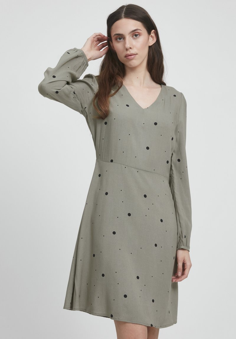 Khakifarbenes Kleid mit V-Ausschnitt und langen Puffärmeln, versehen mit schwarzen Punkten. Aus weichem Stoff mit fließender Silhouette.