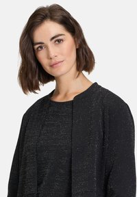 Giacca nera con texture che presenta un leggero luccichio, apertura frontale e scollatura arrotondata, indossata sopra un top abbinato con maniche corte.