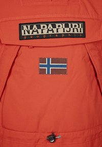 Oranssi kevyt takki, jossa on musta "NAPAPIJRI" -logo, ruskea ja sininen Norjan lipun tarra sekä säädettävä nyöri helmassa.