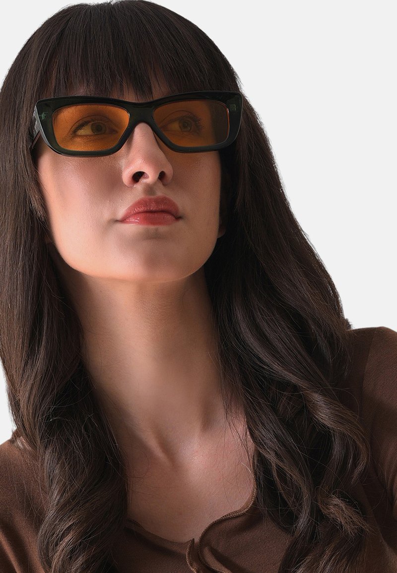 Svarte rektangulære solbriller med amber linser, prydet med et dristigt geometrisk design og en blank finish, båret af en person med langt, mørkt hår.