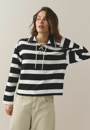 Kobieta o krótkich brązowych włosach, ubrana w czarną i białą koszulę w paski z długim rękawem i sznurowanym kołnierzem, beżowe spodnie, stojąca na jednolitym tle.