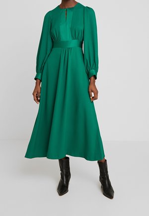 Femme portant une robe verte à manches longues, longue jusqu'aux chevilles avec une taille cintrée et des bottines noires, debout devant un fond uni.