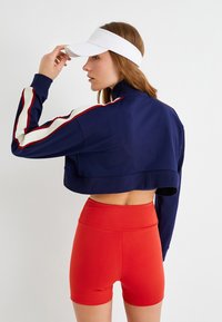 Chaqueta de marinero corta con franjas blancas y rojas en las mangas, combinada con shorts rojos de cintura alta. Visera blanca llevada en la cabeza.