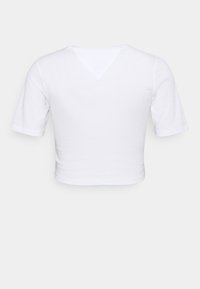 Vit kortärmad cropped skjorta med V-formad sömdetalj vid bakre halsringning, uppvisad mot en enkel bakgrund.
