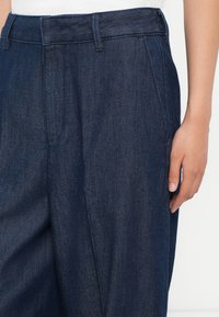 Közelkép sötétkék farmernadrágról, övpántokkal és oldalsó zsebekkel, amelyet egy olyan személy visel, akinek az egyik keze lazán az oldala mellett van.