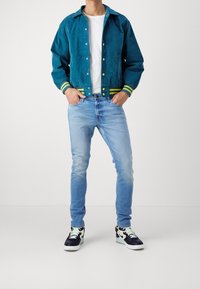 Hombre con chaqueta bomber color teal con puños a rayas, camiseta blanca, jeans ajustados de color azul claro y zapatillas negras y blancas, de pie con las manos en los bolsillos.