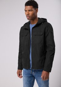 Svart lättviktig jacka med huva, dragkedjestängning, två framfickor och justerbara dragsnören, bärs över en blå skjorta och jeans.