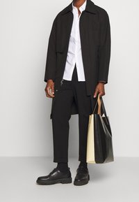 Svart kappa över en vit skjorta, med svarta byxor och skor. En stor tvåfärgad väska, svart och krämfärgad, hålls i ena handen.