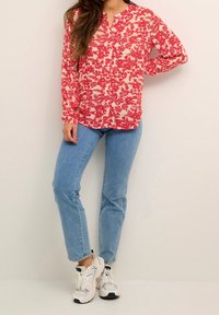 Blusa rossa floreale a maniche lunghe con colletto e chiusura a bottone, abbinata a jeans dritti blu chiaro e sneakers bianche.