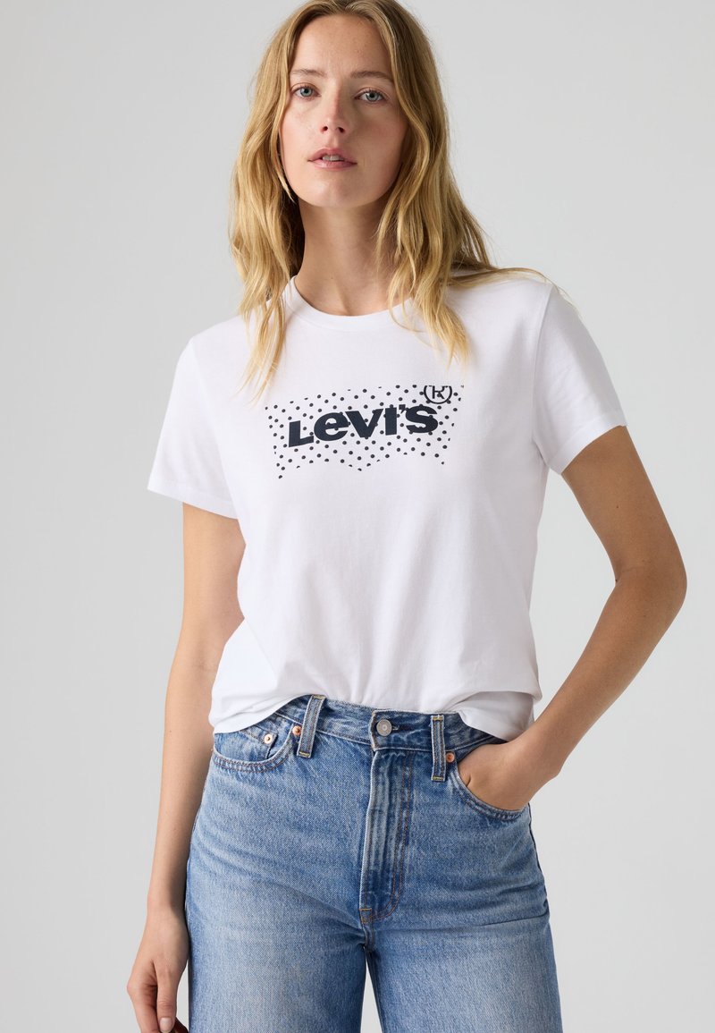 Ξανθιά γυναίκα που φοράει λευκό T-shirt Levi's με μαύρες βούλες και μπλε τζιν με ψηλή μέση, στέκεται με το ένα χέρι στην τσέπη μπροστά σε γκρι φόντο.