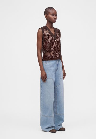 Femme portant un haut marron sans manches à paillettes et un jean bleu clair à jambes larges, debout sur fond blanc.
