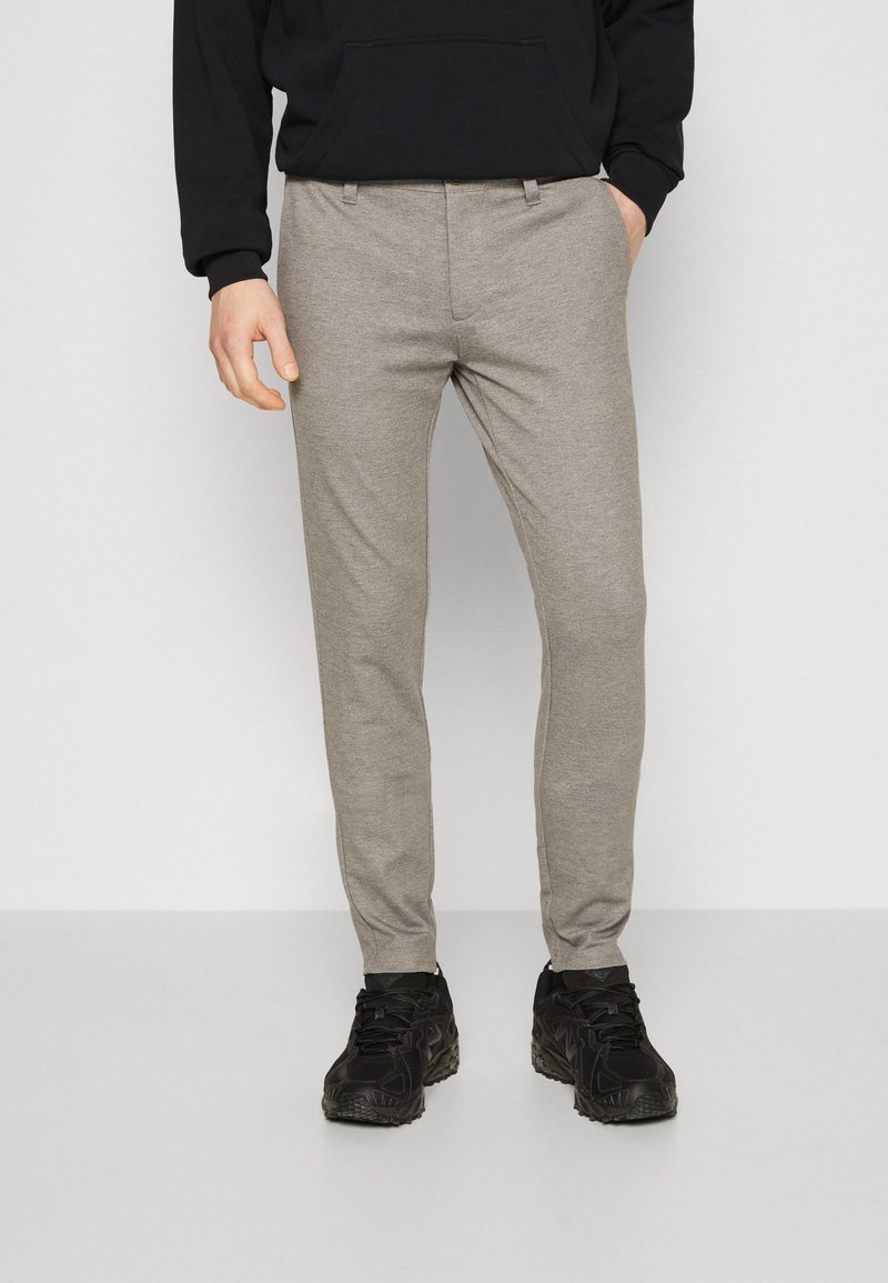 Homme portant un pantalon gris coupe slim et des baskets noires, debout avec une main dans la poche devant un fond blanc uni.