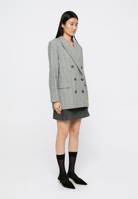 Femme se tenant dans un blazer croisé à rayures grises, une jupe plissée noire, des chaussettes noires transparentes et des escarpins noirs pointus sur un fond blanc.