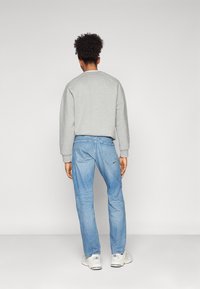 Lys grå sweatshirt med rund hals, kombinert med lyseblå jeans. Modellen har på seg hvite joggesko, sett fra bakgrunnen mot en ensfarget bakgrunn.