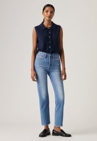 Γυναίκα στέκεται φορώντας μπλε σκούρο αμάνικο πουκάμισο με κουμπιά, ανοιχτό μπλε ψηλόμεσο τζιν και μαύρα μοκασίνια μπροστά σε απλό φόντο.