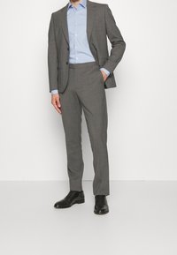 Grijs tailored pak met een enkelrijige jas en broeken met een platte voorkant. Lichtblauw overhemd met knopen en zwarte leren nette schoenen.