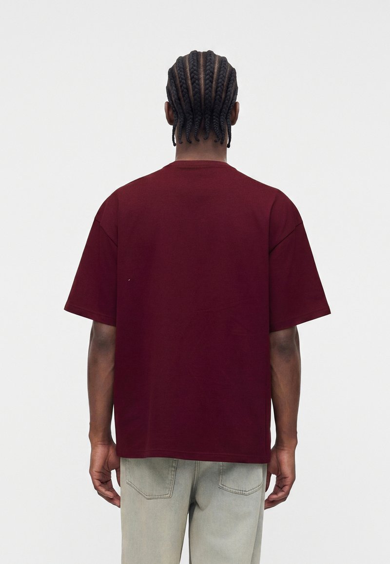 Mann mit geflochtenem Haar trägt ein locker sitzendes dunkelrotes kurzärmeliges T-Shirt und hellgraue Hose und steht mit dem Rücken zum Betrachter.