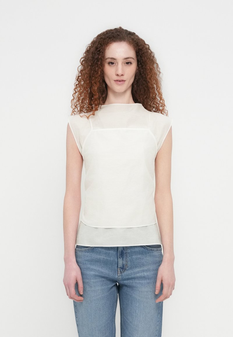 Γυναίκα με καστανά σγουρά μαλλιά, φοράει λευκό αμάνικο τοπ με στρώσεις και μπλε τζιν, στέκεται μπροστά σε απλό λευκό φόντο.