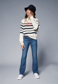 Vit och marinrandig sweatshirttopp med röd logotyp, kombinerad med blå utsvängda jeans och vita sneakers; kompletterad med en marinblå bucket-hatt.