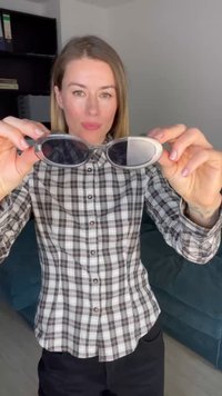 Gafas de sol redondas de color plateado con lentes oscuros y un marco liso, sostenidas frente a una persona que lleva una camisa de cuadros.