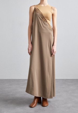 Mouwloze bruine maxi-jurk met kantafwerking, gemaakt van een gladde stof. Draagt over een beige top, gecombineerd met bruine haklaarzen.