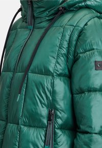 Zelená péřová bunda s lesklou, prošívanou texturou. Obsahuje kapuci, nastavitelné šňůrky a zipovou kapsu na boku.