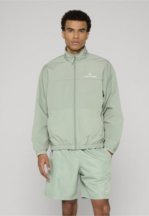 Mężczyzna w jasnorozowym rozpinanym dresie i dopasowanych szortach stoi na tle szarego, jednolitego tła, z jedną ręką w kieszeni.