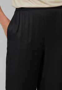 Zwarte, lichte broek met een elastische tailleband, voorzien van zijzakken en een gladde, iets glanzende textuur.