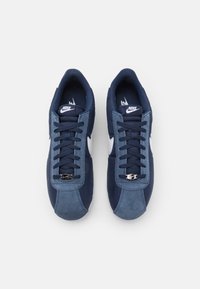 Par tenisica u tamnoplavoj nijansi marke Nike s bijelim swooshom, odsuđenim prednjim dijelom od svežnja i crnim vezicama, snimljenih odozgo na bijeloj pozadini.