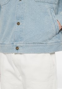 Ljusblå denimjacka med knäppning framtill, bröstfickor och en mjuk textur, ihop med vita avslappnade byxor.