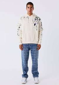 Crème hoodie met zwart en paars spetterontwerp, kangoeroezak; gecombineerd met lichtblauwe jeans met witte logotext.