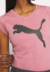 Różowa koszulka sportowa z dekoltem w serek, z dużym czarnym logo Puma. Materiał wydaje się miękki z delikatną teksturą. Krótkie rękawy.